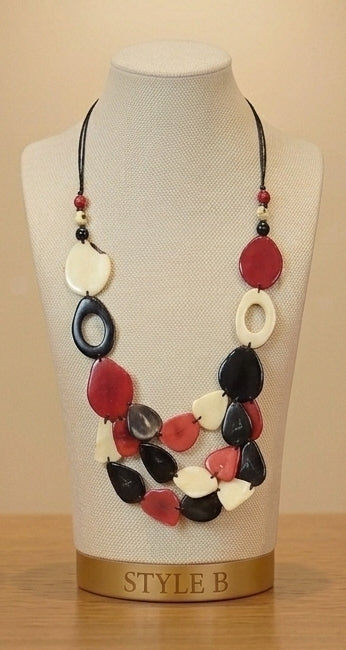 A statement necklace with irregular red, black, and white tagua beads on a black cord, displayed on a cream fabric mannequin bust with a gold base labeled 'STYLE B'.