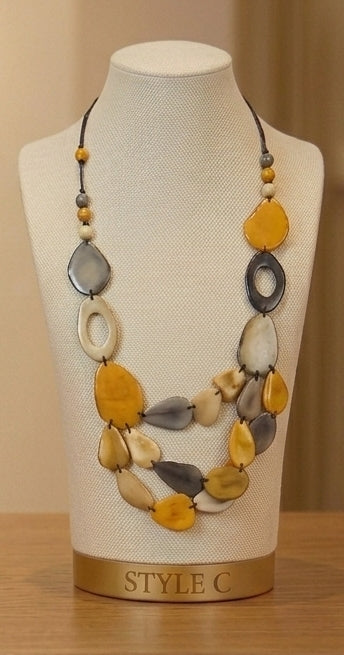 A multi-strand statement necklace with organic-shaped beads in yellow, gray, beige, and white, displayed on a cream fabric mannequin bust labeled 'STYLE C'.