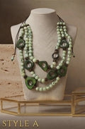 A multi-strand statement necklace displayed on a beige fabric mannequin bust. The necklace features light sage green beads, dark green tagua oval accents, and dark brown cord details.
