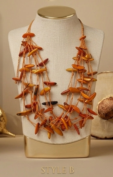 A multi-strand statement necklace made of elongated amber-orange and brown shell-like pieces on tan cords, displayed on a cream fabric jewelry bust with a gold neck cap and base. The bust is centered against a warm beige background with decorative objects partially visible at the sides.