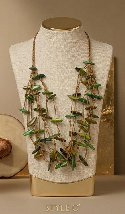 A multi-strand statement necklace with green stone-like discs and gold-toned cords, displayed on a cream fabric-covered jewelry bust with a gold base. The bust is set against a warm beige background with decorative props.