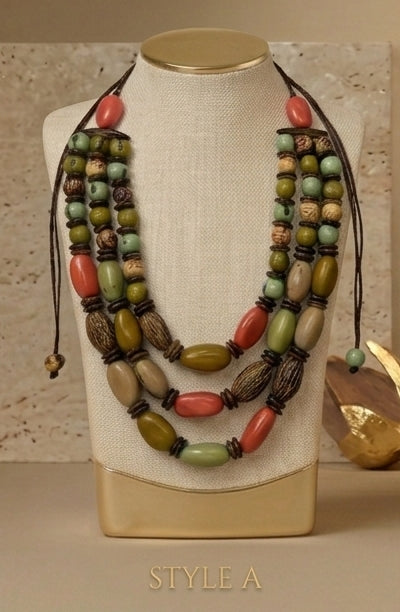 A multi-strand statement necklace displayed on a cream fabric mannequin bust. The necklace features a mix of olive green, terracotta red, brown, and beige beads with dark brown cord ties.