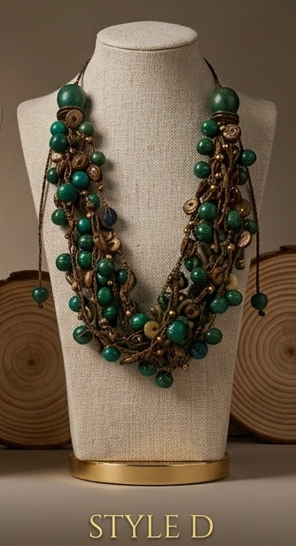 A multi-strand statement necklace with green beads and bronze-toned metal details, displayed on a beige fabric jewelry mannequin bust with wood slice props in the background.