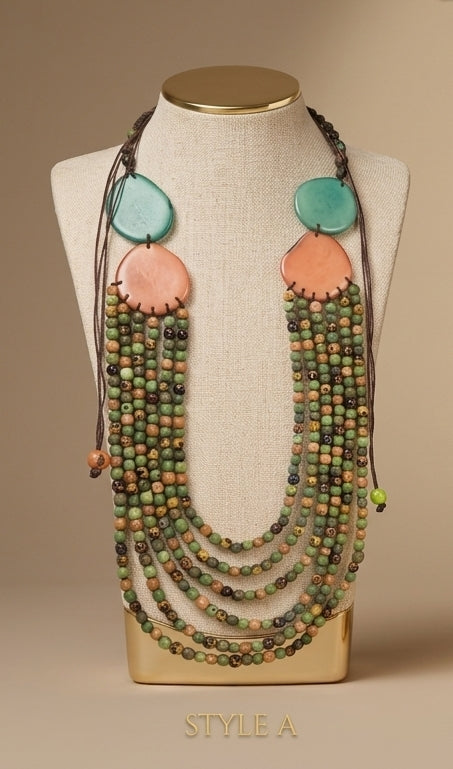 A multi-strand statement necklace displayed on a beige fabric mannequin bust. The necklace features multiple strands of green and brown beads, large peach-pink and turquoise disc pendants, and dark brown cords.