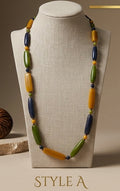 A long beaded necklace displayed on a beige fabric mannequin bust. The necklace features alternating large olive green, mustard yellow, and dark navy blue beads with small yellow spacer beads.