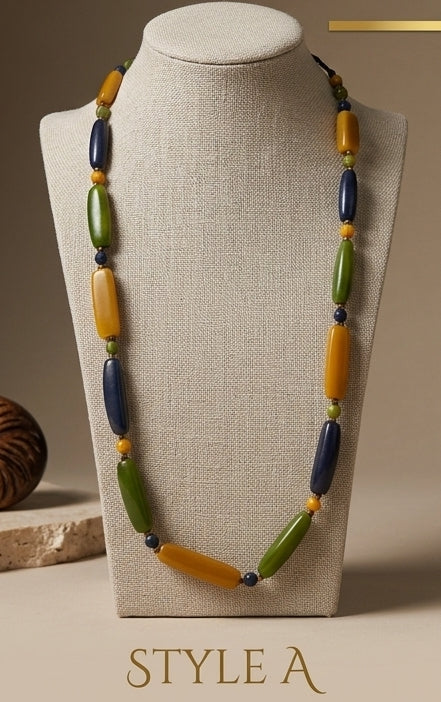 A long beaded necklace displayed on a beige fabric mannequin bust. The necklace features alternating large olive green, mustard yellow, and dark navy blue beads with small yellow spacer beads.