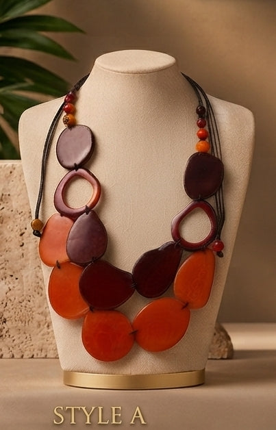 A statement necklace displayed on a cream jewelry bust. The necklace features large, organic-shaped beads in shades of orange, terracotta, and deep burgundy, strung on black cords with small orange and red beads.