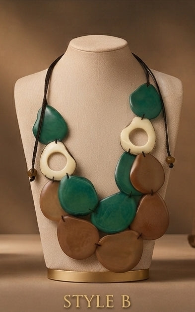 A statement necklace displayed on a beige jewelry bust against a warm brown gradient background. The necklace features large, organic-shaped beads in teal green, cream white, and earthy brown, suspended on a black cord with small gold-toned beads.
