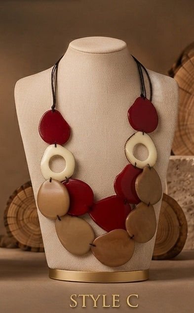 A statement necklace displayed on a cream-colored mannequin bust, featuring large organic-shaped beads in red, cream, and tan tones. The necklace is suspended from black cords and is set against a warm, rustic background with wood slices.