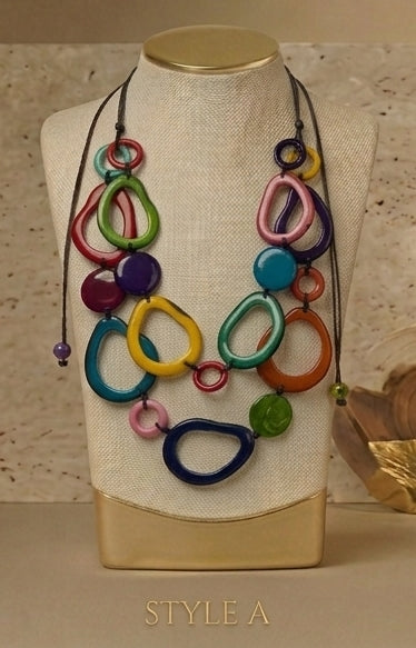A multicolor statement necklace with organic-shaped beads and layered rings on a black cord, displayed on a beige fabric mannequin bust. The necklace features shades of blue, green, pink, yellow, orange, purple, and red.