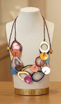 A multicolor statement necklace displayed on a cream fabric jewelry bust. The necklace features a mix of colorful circular, oval, and ring-shaped beads in shades of orange, yellow, pink, blue, purple, and white, suspended from dark brown cords.