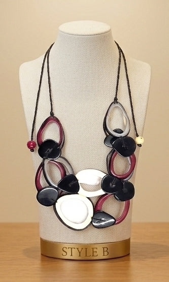 A statement necklace with layered black, white, and burgundy oval and circular elements on a cream mannequin bust, displayed on a wooden surface against a beige background.