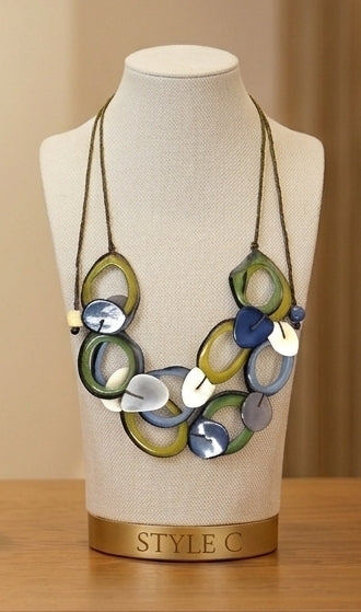 A multicolor statement necklace with overlapping circular and oval links in olive green, blue, gray, and white, displayed on a cream fabric jewelry bust labeled 'STYLE C'.