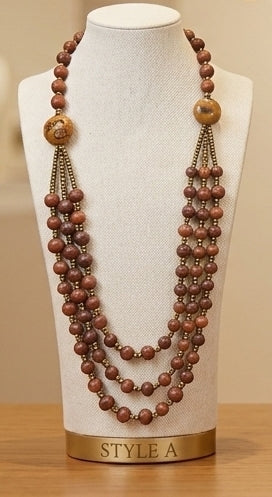 A multi-strand beaded necklace displayed on a cream fabric mannequin bust. The necklace features multiple strands of medium brown wooden beads, small metallic bronze spacer beads, and two larger round wooden pendant-like beads near the top.