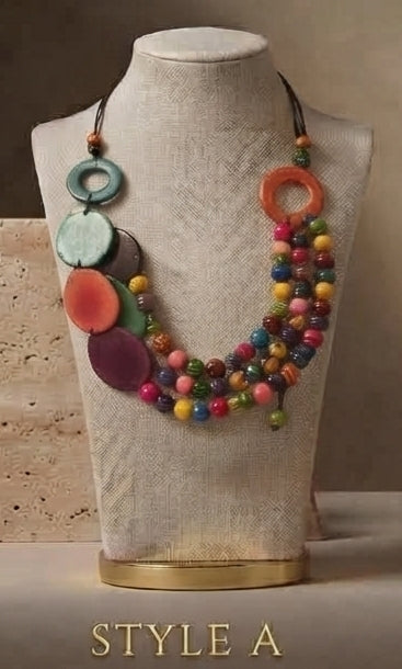 A multicolor statement necklace displayed on a beige fabric mannequin bust. The necklace features large circular disc charms in orange, green, purple, and pink, alongside strands of small multicolored beads.