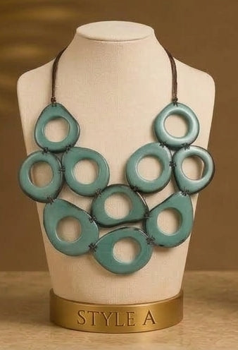 A statement bib necklace with multiple layered turquoise-green circular cutout pendants on a dark cord, displayed on a cream jewelry bust labeled 'STYLE A' against a warm brown background.