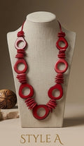 A red beaded necklace with large circular and spiral bead motifs displayed on a beige fabric-covered jewelry bust. The bust is set against a warm beige background, with a small decorative object partially visible on the left and the text 'STYLE A' at the bottom.