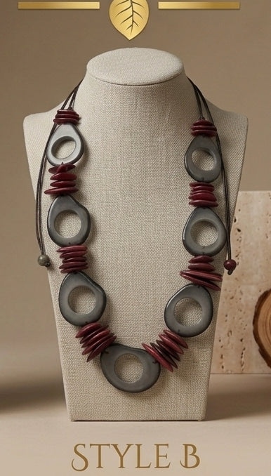 A statement necklace displayed on a beige fabric-covered jewelry bust. The necklace features large, dark gray circular disc beads and clusters of dark red disc beads on a black cord.