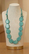 A turquoise beaded necklace displayed on a cream fabric mannequin bust with a gold base labeled 'STYLE A', set against a warm beige background.