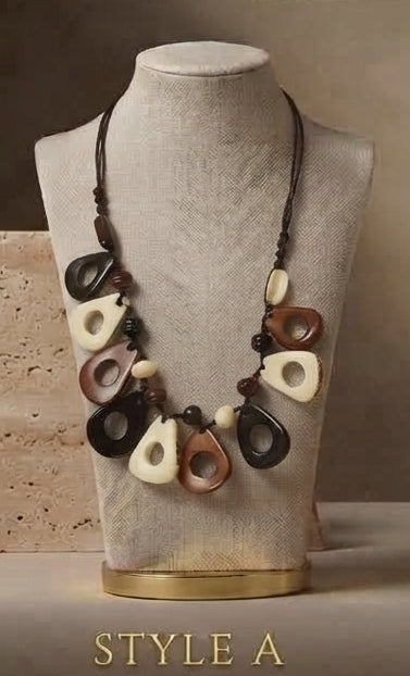 A statement necklace with organic-shaped beads in black, brown, and cream, displayed on a textured beige mannequin bust with a gold base. The text 'STYLE A' is printed at the bottom.