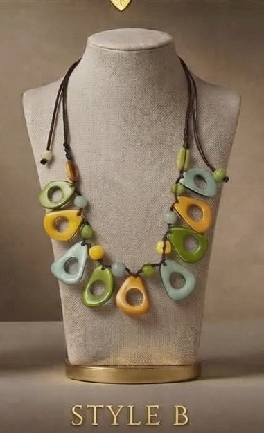A bohemian-style multi-color pendant necklace displayed on a beige fabric mannequin bust. The necklace features organic-shaped beads in yellow, green, and light blue on dark brown cords, with the text 'STYLE B' printed at the bottom.