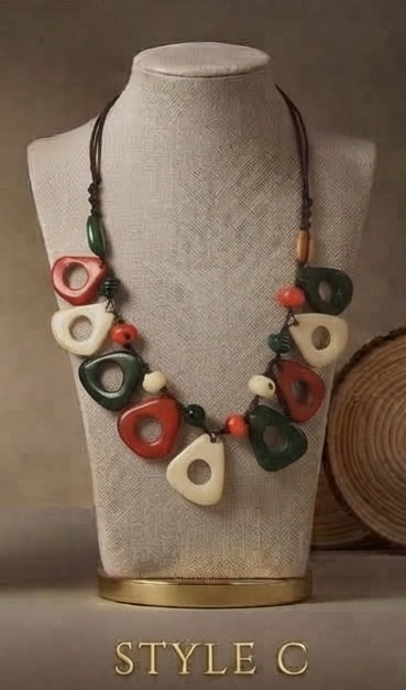A statement necklace with chunky, organic-shaped beads in red, green, and cream, displayed on a beige fabric jewelry bust. The necklace is suspended from a dark cord and features small orange and white beads as accents.