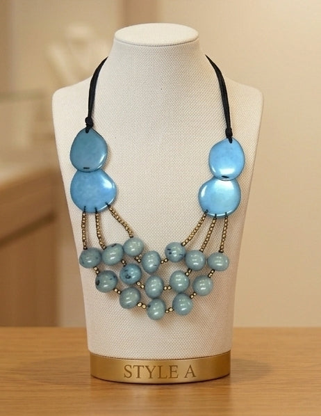 A statement necklace displayed on a cream fabric mannequin bust labeled 'STYLE A'. The necklace features three large light blue disc-shaped beads on the left side, three matching light blue disc-shaped beads on the right side, and multiple strands of smaller light blue marbled beads hanging from gold-toned chains.