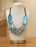 A statement necklace displayed on a cream fabric mannequin bust labeled 'STYLE A'. The necklace features three large light blue disc-shaped beads on the left side, three matching light blue disc-shaped beads on the right side, and multiple strands of smaller light blue marbled beads hanging from gold-toned chains.