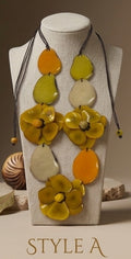 A statement necklace displayed on a beige fabric mannequin bust. The necklace features large, organic-shaped beads in yellow, orange, and off-white, accented by two prominent yellow flower charms with textured centers, all suspended from dark cords.
