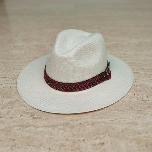 A white woven fedora-style hat with a dark red and black herringbone patterned band, resting on a light beige stone surface. Ecuadorian Toquilla Straw Panama Hat.