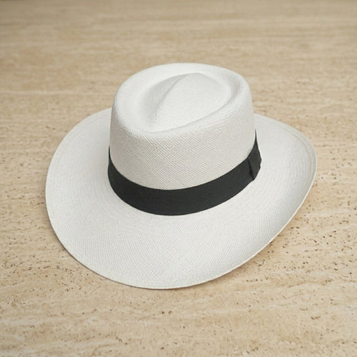 A white woven fedora-style hat with a black ribbon band, photographed from above on a light beige textured surface. Ecuadorian Toquilla Straw Hat Paso Fino white.