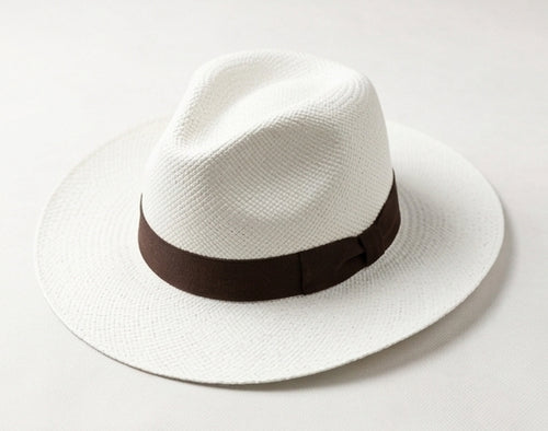 A white woven fedora hat with a dark brown band around the crown, photographed on a clean light gray background. Ecuadorian Toquilla Straw Panama Hat.
