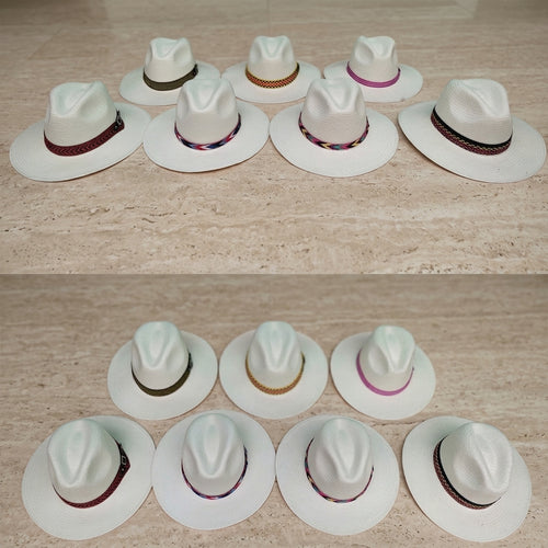 Nine white woven fedora-style hats with multicolored patterned bands are arranged in two rows on a light beige stone surface. Ecuadorian Toquilla Straw Panama Hats.