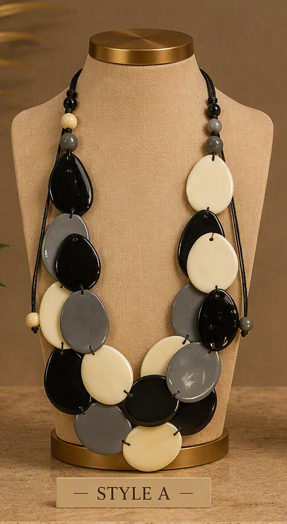 A statement necklace with large oval disc beads in black, cream, and gray, displayed on a beige fabric jewelry bust with a gold-toned base. A small label at the bottom reads 'STYLE A'.