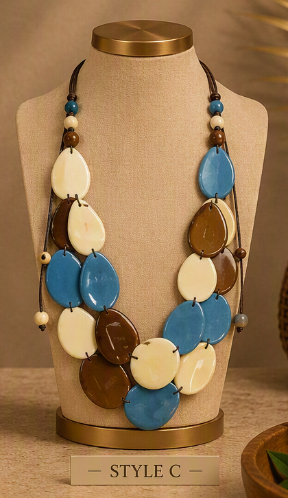 A multi-strand statement necklace displayed on a beige fabric jewelry mannequin bust. The necklace features large, organic-shaped disc elements in blue, cream, and brown, suspended on dark cords with small accent beads.