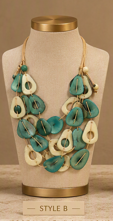 A multi-strand statement necklace displayed on a beige fabric jewelry bust. The necklace features alternating large, organic-shaped beads in teal green and cream, suspended on a tan cord.