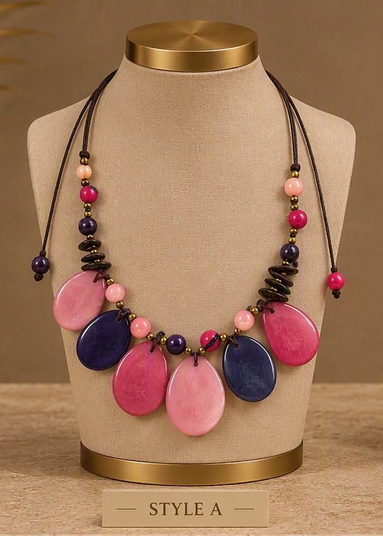 A statement necklace displayed on a beige fabric mannequin bust with a gold-toned collar. The necklace features large, teardrop-shaped beads in pink and navy blue, accented with smaller pink and dark purple beads, and black disc-like beads on black cords.