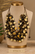 A multi-strand statement necklace displayed on a beige fabric mannequin bust with gold-toned caps. The necklace features layered dark brown cords adorned with round, textured shell-like beads in cream, black, and brown mottled patterns.