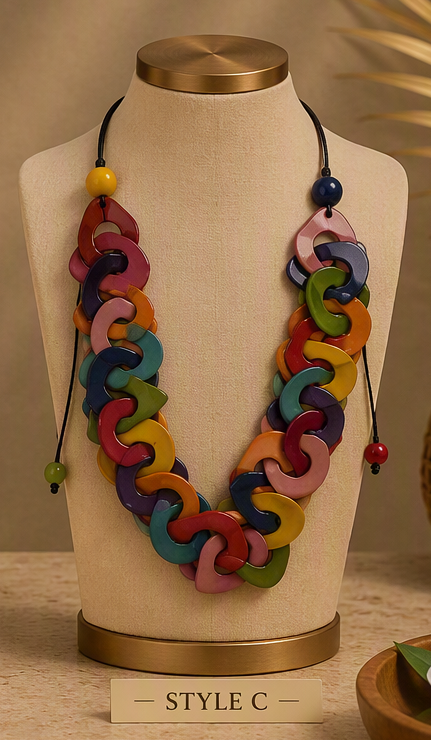 A multicolor wooden link necklace displayed on a beige fabric mannequin bust with a gold-toned base. The necklace features interlocking curved wooden links in shades of pink, purple, blue, green, yellow, and orange, suspended from black cords with decorative beads, Ecuadorian tagua necklace.