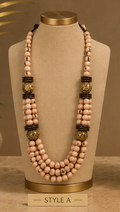 A multi-strand beaded necklace displayed on a beige jewelry bust. The necklace features multiple strands of light pinkish-beige beads, accented with dark brown textured spacer beads and two large round gold-toned beads with black detailing.