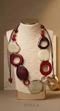 A multi-strand statement necklace displayed on a textured cream fabric mannequin bust. The necklace features dark brown cords, red and cream-colored tagua disc pendants, and small red beads.