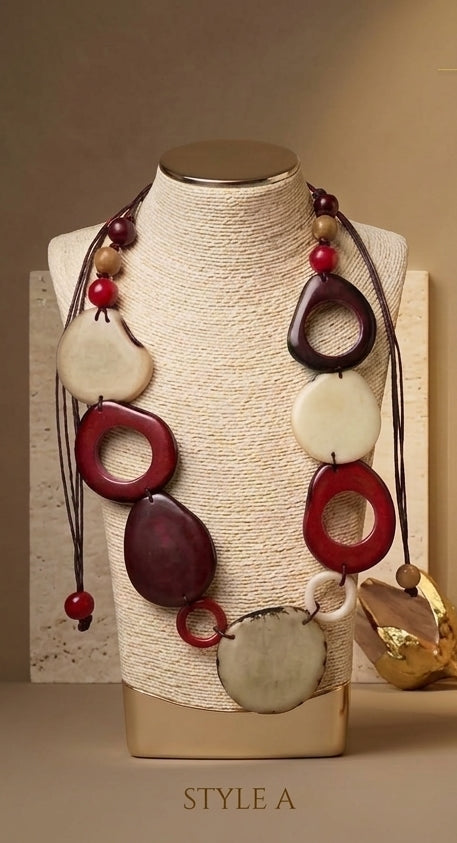 A multi-strand statement necklace displayed on a textured cream fabric mannequin bust. The necklace features dark brown cords, red and cream-colored tagua disc pendants, and small red beads.
