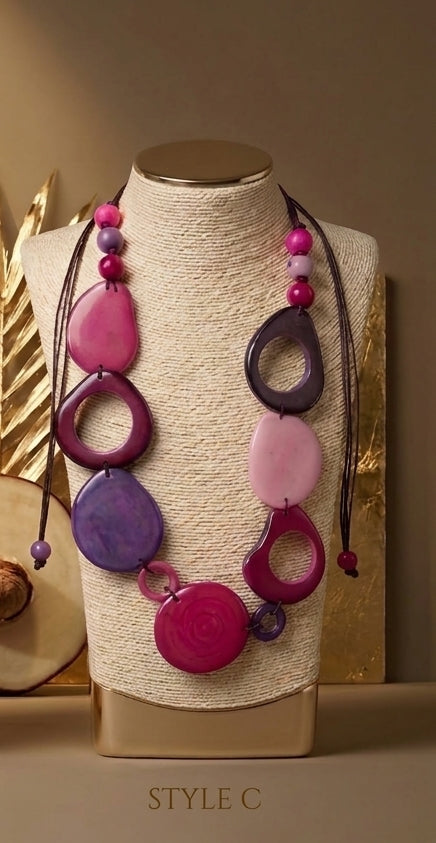 A bohemian-style multi-colored beaded necklace displayed on a textured cream-colored mannequin bust. The necklace features large, irregular tagua beads in shades of magenta, purple, and pink, strung on dark cords.