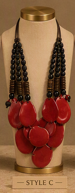 A statement necklace displayed on a beige fabric mannequin bust with a gold-toned top and base. The necklace features multiple strands of glossy black beads with small metallic spacer details, paired with large, glossy red teardrop-shaped beads at the center.
